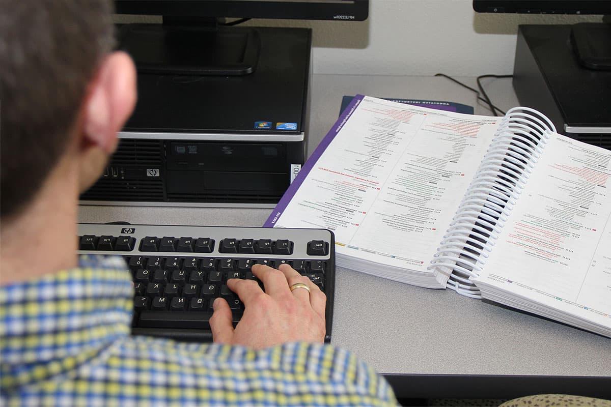 一个人坐在电脑前, typing on the keyboard, 他们旁边的桌子上放着一本医学编码信息的书